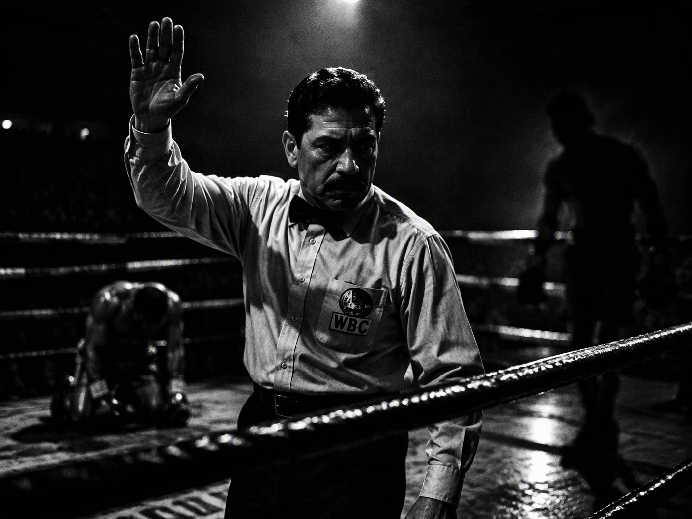 Black-and-white image of boxing referee Russell Mora illuminated by a single overhead light in a shadow-filled ring. A boxer kneels in the background as ropes and shadows crisscross the scene, symbolizing the tension and controversy of a disputed call.