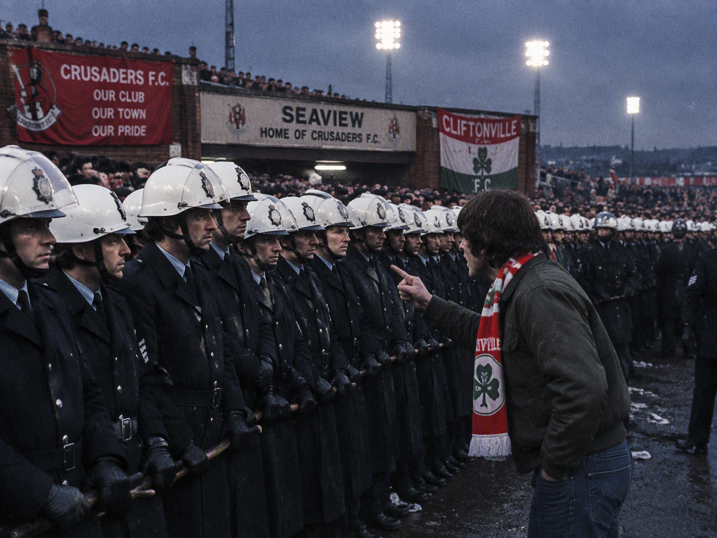 A late 1970s color photograph of a large line of police officers in helmets standing in front of Seaview football ground under stadium lights, with a lone Cliftonville supporter in a red scarf facing them, capturing the tense atmosphere of the 1979 North Belfast derby.