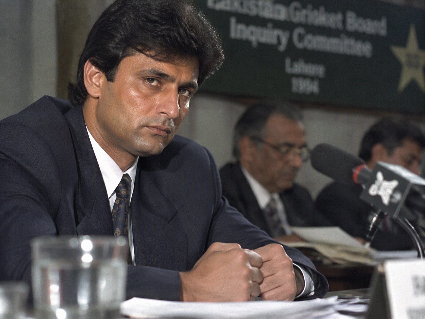 Portrait of Salim Malik at the Qayyum Commission hearing in 1999, illuminated by harsh lighting, his hands clenched on the table conveying tension amid the match-fixing allegations.